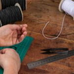 A person measuring their wrist with black and white elastic cords, sitting at a wooden table. The table also has spools of black and white elastic cords, a green cutting mat, a metal ruler, and a pair of black scissors.