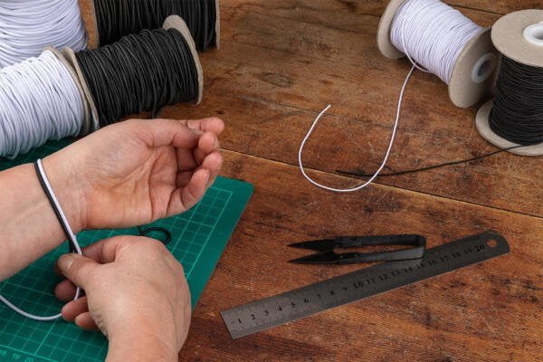 A person measuring their wrist with black and white elastic cords, sitting at a wooden table. The table also has spools of black and white elastic cords, a green cutting mat, a metal ruler, and a pair of black scissors.