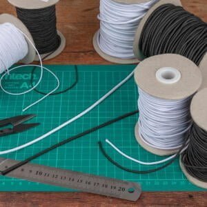 Multiple spools of elastic cord in black and white are arranged on a cutting mat. Scissors, a metal ruler, and a cable tie are also seen on the mat. The cutting mat is green and features measurement markings. The background is a wooden table.