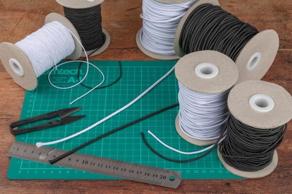 Multiple spools of elastic cord in black and white are arranged on a cutting mat. Scissors, a metal ruler, and a cable tie are also seen on the mat. The cutting mat is green and features measurement markings. The background is a wooden table.