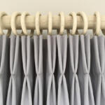 A close-up of light gray curtain panels hanging from a wooden rod with large, circular, white rings. The curtains are pleated and evenly spaced, showing the top section in detail. The background features a neutral-colored wall.