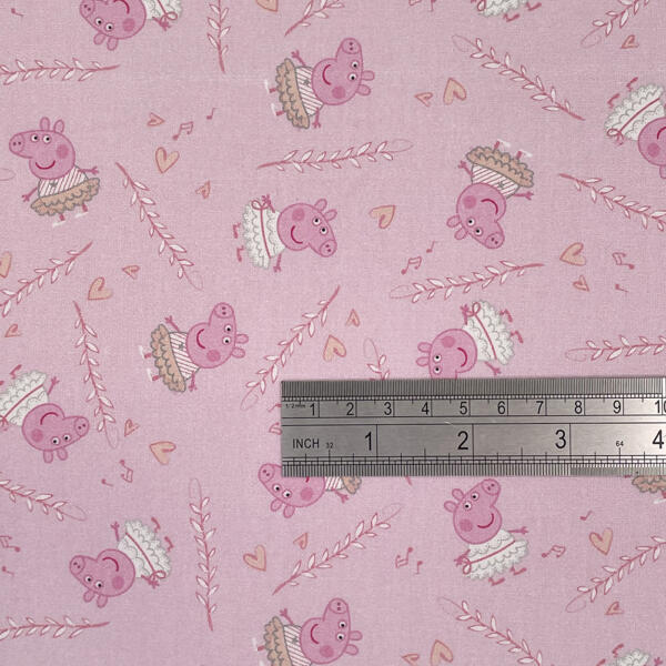 A close-up of a fabric with a pink background, featuring a pattern of cartoon pigs dressed in tutus and dancing. The fabric also has musical notes, hearts, and leafy branches. A ruler at the bottom shows measurements in inches and centimeters for scale.