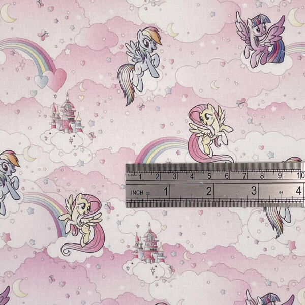 A pink fabric features colorful illustrations of flying ponies, castles, rainbows, and clouds in a whimsical design. A ruler placed on the fabric shows measurements in inches and centimeters, suggesting scale for the pattern.