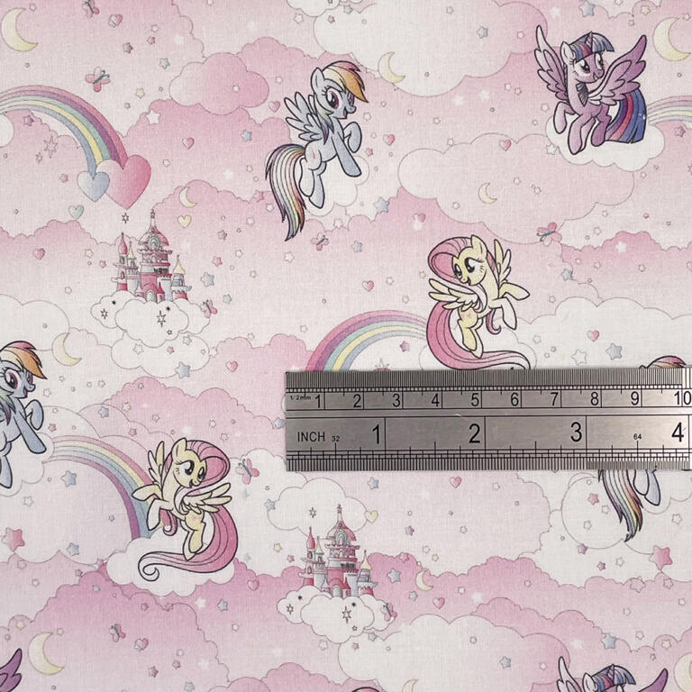 A pink fabric features colorful illustrations of flying ponies, castles, rainbows, and clouds in a whimsical design. A ruler placed on the fabric shows measurements in inches and centimeters, suggesting scale for the pattern.