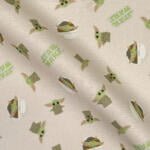Fabric with repeated "Star Wars" logos and illustrations of an alien-like character with large ears, a green face, and a beige robe. The characters are depicted in various poses and settings, including peeking out of a pod and waving.