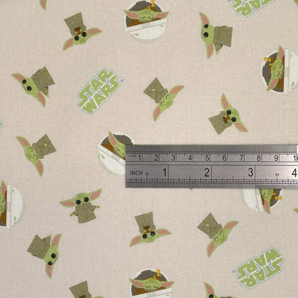 Fabric featuring a playful Baby Yoda (Grogu) pattern from the Star Wars series, shown with a silver ruler for scale. Baby Yoda is depicted in various poses against a light background, with "Star Wars" logos intermittently placed in the design.
