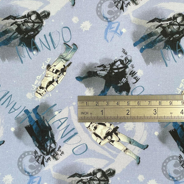 A fabric with a Star Wars theme featuring illustrations of characters in armor, primarily in shades of gray and blue. The words "Mando" and "This is the Way" are visible. A wooden ruler is placed on the fabric for scale, showing inches and centimeters.