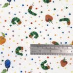 A fabric featuring a colorful print of the Very Hungry Caterpillar, fruits (oranges, strawberries, plums), and multicolored polka dots on a white background. A ruler showing inches and centimeters is placed on the fabric for scale.