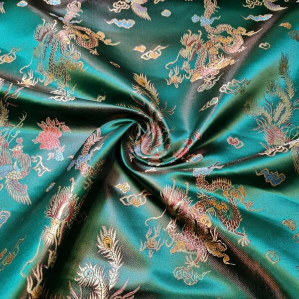 A close-up of a fabric with an intricate, colorful dragon and floral pattern. The fabric is a rich, glossy teal color, and the pattern includes gold, red, and other metallic threads. The fabric is gathered in the center into a swirl shape.