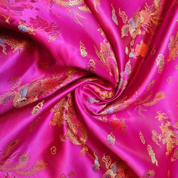 A close-up image of vibrant pink silk fabric adorned with intricate gold, red, and blue embroidered designs. The fabric is gathered in the center, forming a swirl pattern, showcasing the detailed and colorful embroidery.