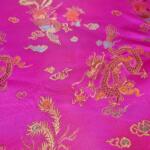 A rich, magenta fabric adorned with intricate gold and blue embroidery of dragons and other traditional Asian motifs. The detailed designs create a luxurious and vibrant pattern across the satin-like material, showcasing cultural artistry.