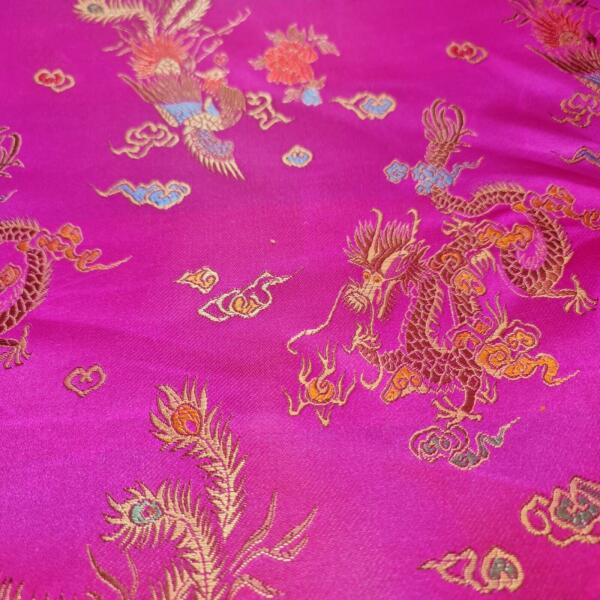 A rich, magenta fabric adorned with intricate gold and blue embroidery of dragons and other traditional Asian motifs. The detailed designs create a luxurious and vibrant pattern across the satin-like material, showcasing cultural artistry.