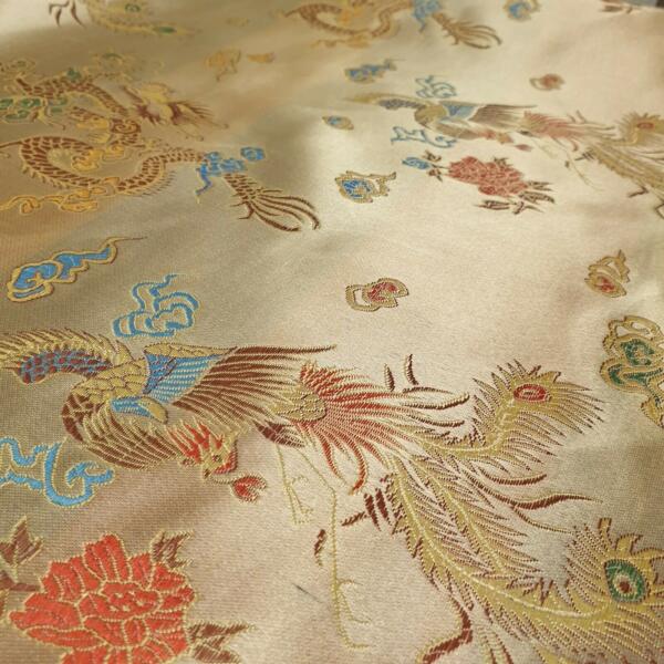 Close-up of an intricate fabric with a beige background featuring colorful, embroidered patterns of dragons, phoenixes, clouds, and flowers in red, blue, green, and yellow. The detailed design showcases traditional Asian artistic elements.