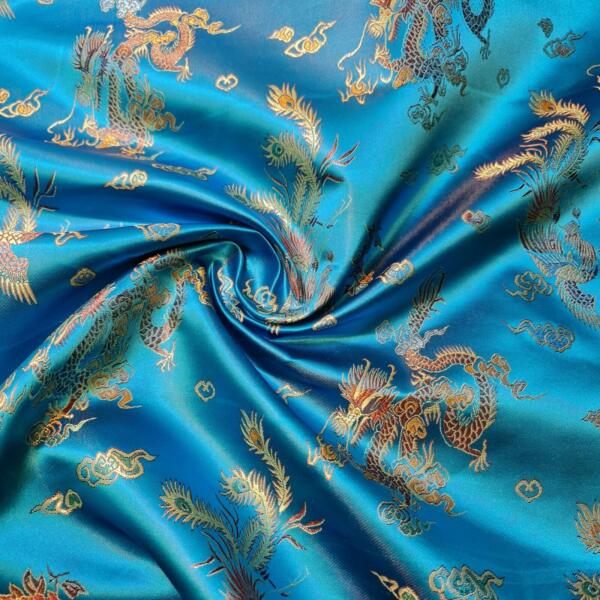 A close-up of vibrant blue silk fabric featuring an intricate pattern of gold embroidered dragons and flora. The material has luxurious texture, folds in elegant waves, and catches light, highlighting the detailed and decorative design.