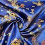 A close-up of a luxurious, deep blue silk fabric adorned with intricate gold and multicolored embroidery. The pattern features ornate designs, including phoenixes and floral motifs, with the fabric gathered into a central, swirling knot.