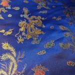 A close-up of a rich blue fabric adorned with vibrant, intricate embroidery. The design features golden dragons, multicolored flowers, and peacock feathers. The fabric appears to be silky, with the detailed patterns reflecting light to create a shimmering effect.