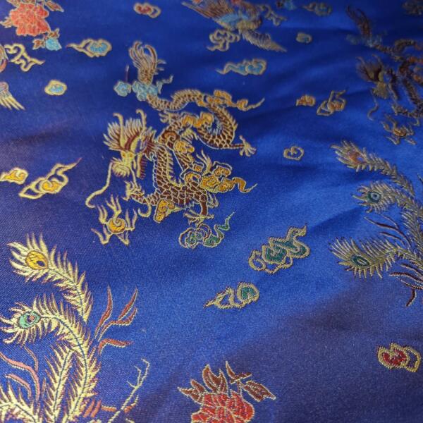 A close-up of a rich blue fabric adorned with vibrant, intricate embroidery. The design features golden dragons, multicolored flowers, and peacock feathers. The fabric appears to be silky, with the detailed patterns reflecting light to create a shimmering effect.