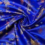 A close-up image of a vibrant royal blue silk fabric with intricate gold, red, and turquoise embroidery, featuring delicate floral and mythical bird patterns. The fabric is gently twisted in the center, showcasing the luxurious texture and detailed design.