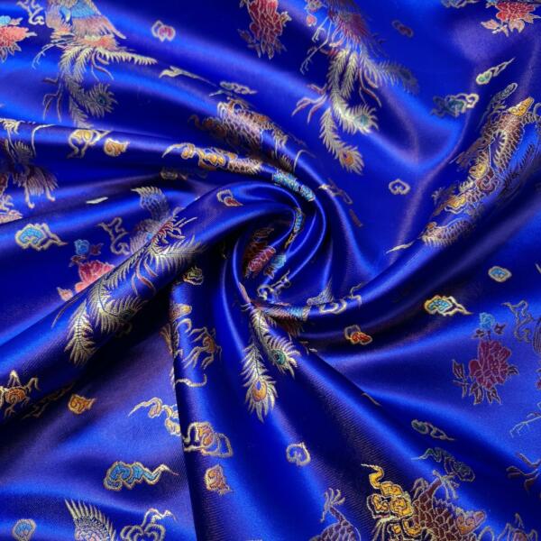A close-up image of a vibrant royal blue silk fabric with intricate gold, red, and turquoise embroidery, featuring delicate floral and mythical bird patterns. The fabric is gently twisted in the center, showcasing the luxurious texture and detailed design.
