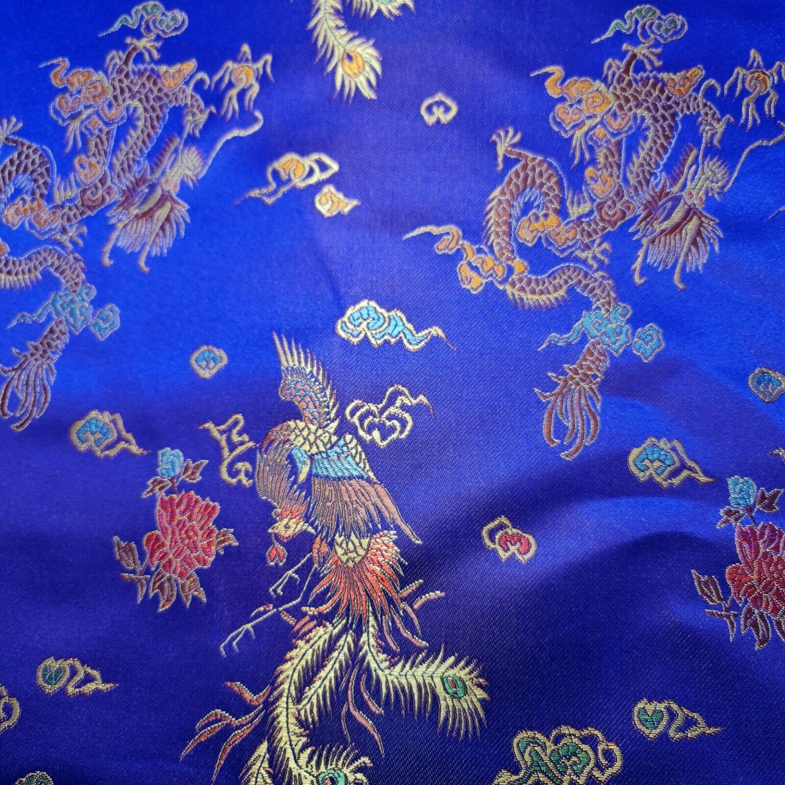 A piece of blue silk fabric featuring intricate, multicolored embroidery of mythical Eastern dragons and phoenixes among clouds and flowers. The vibrant design includes gold, red, green, and blue threads, creating a rich and detailed pattern against the smooth background.