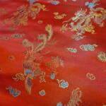 A close-up of red embroidered fabric featuring intricate designs of dragons and phoenixes. The patterns include gold, blue, and white thread, creating a rich, textured appearance that highlights the detailed mythical creatures against the red background.