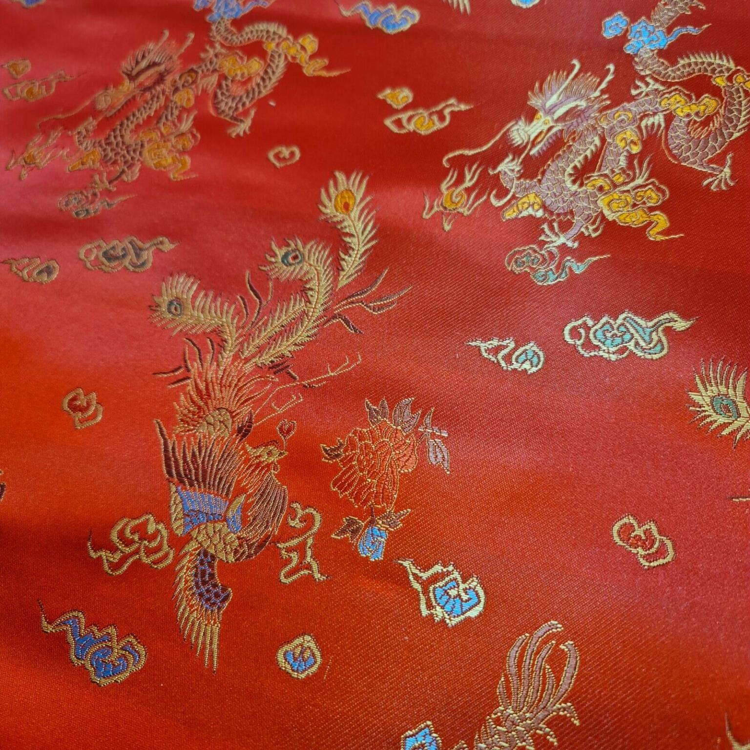 A close-up of red embroidered fabric featuring intricate designs of dragons and phoenixes. The patterns include gold, blue, and white thread, creating a rich, textured appearance that highlights the detailed mythical creatures against the red background.
