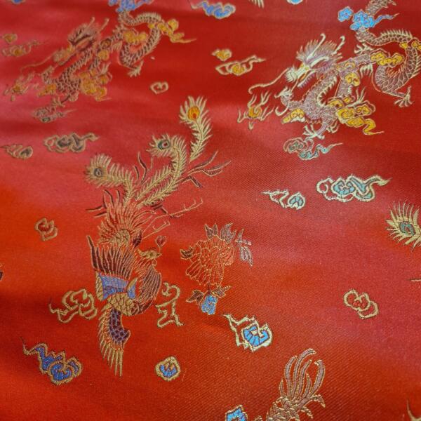 A close-up of red embroidered fabric featuring intricate designs of dragons and phoenixes. The patterns include gold, blue, and white thread, creating a rich, textured appearance that highlights the detailed mythical creatures against the red background.