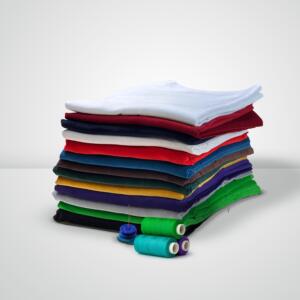 A stack of neatly folded, multicolored fabric pieces is placed on a surface. In front of the stack are three spools of thread in green, blue, and white with a blue button and a sewing needle threaded with green thread.