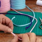 Close-up of hands crafting with colorful ropes on a green cutting mat. Scissors, a metal ruler, and spools of pink, blue, and purple rope are visible. A spool of white thread and scattered golden pins are also seen on a wooden surface.