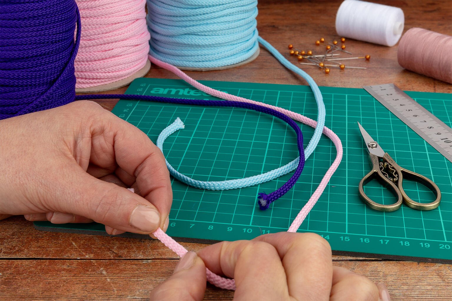 Close-up of hands crafting with colorful ropes on a green cutting mat. Scissors, a metal ruler, and spools of pink, blue, and purple rope are visible. A spool of white thread and scattered golden pins are also seen on a wooden surface.