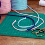 Scissors, a ruler, and a cutting mat are arranged on a wooden surface. Coiled ropes in pink, blue, purple, white thread, and several pins are also present, ready for crafting.