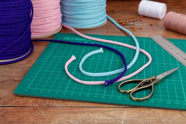 Scissors, a ruler, and a cutting mat are arranged on a wooden surface. Coiled ropes in pink, blue, purple, white thread, and several pins are also present, ready for crafting.