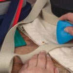 A person's hands using a blue tailor's chalk to mark a beige fabric that's pinned on a wooden surface. Various rolls of colorful woven tapes and a green cutting mat are visible in the background. The scene is part of a sewing or crafting project.