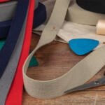 A variety of fabric strips in red, blue, gray, and beige are spread out on a wooden table. A pair of scissors, spools of thread, a blue fabric chalk, and a green cutting mat are also visible, suggesting a sewing or crafting setup.
