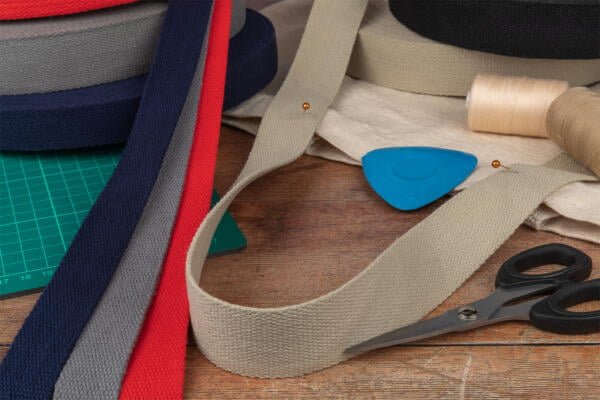 A variety of fabric strips in red, blue, gray, and beige are spread out on a wooden table. A pair of scissors, spools of thread, a blue fabric chalk, and a green cutting mat are also visible, suggesting a sewing or crafting setup.
