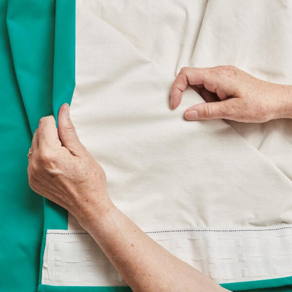Close-up of two hands examining the hem of a fabric with contrasting colors. The left hand is holding the turquoise part of the fabric, while the right hand is lightly touching the white section. The fabric is laid out flat, showing the stitching and hem details.