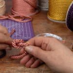 A pair of hands tying a decorative knot with shiny, twisted cords. Various spools of different colored cords are on the table, along with a purple grid cutting mat, a rolled-up measuring tape, and a box of orange sewing pins.