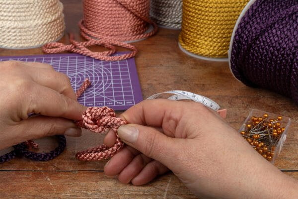 A pair of hands tying a decorative knot with shiny, twisted cords. Various spools of different colored cords are on the table, along with a purple grid cutting mat, a rolled-up measuring tape, and a box of orange sewing pins.