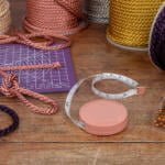 Various colorful cords, a purple cutting mat, measuring tape, and a box of gold-headed pins are arranged on a wooden table. The cords are in different colors including cream, copper, pink, purple, and dark purple. Some cords are wrapped in spools, and one is knotted.