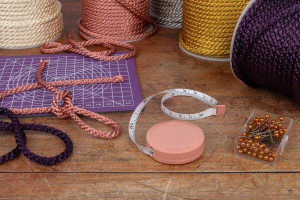Various colorful cords, a purple cutting mat, measuring tape, and a box of gold-headed pins are arranged on a wooden table. The cords are in different colors including cream, copper, pink, purple, and dark purple. Some cords are wrapped in spools, and one is knotted.