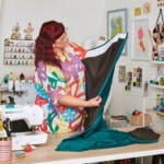 A person with red hair and glasses, wearing a colorful shirt, works in a bright and cluttered sewing room. They are holding and examining a piece of fabric next to a sewing machine. Shelves, lockers, and craft supplies are visible in the background.