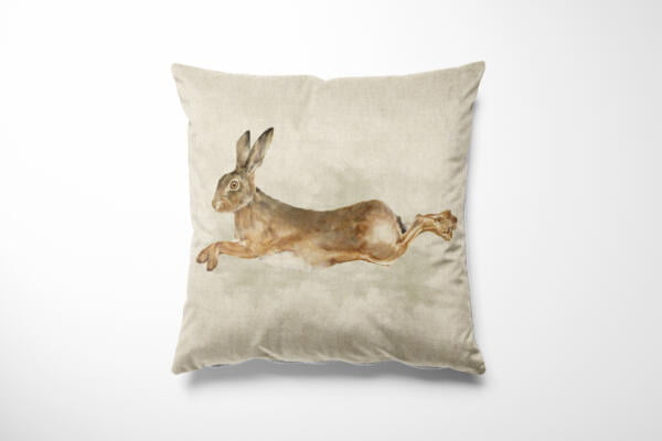 A beige pillow with a realistic painting of a leaping rabbit in the center. The rabbit is depicted mid-jump, with brown and white fur, long ears, and expressive eyes, set against the plain background of the pillow. The overall design has a natural and serene feel.