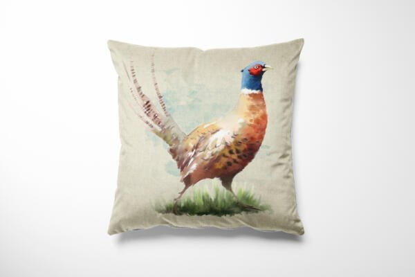 A square pillow with a beige cover features a watercolor illustration of a pheasant. The bird, detailed with vibrant colors including blue, red, and brown, stands on grass with its long tail feathers extended gracefully. The background is a subtle blend of blue and green shades.
