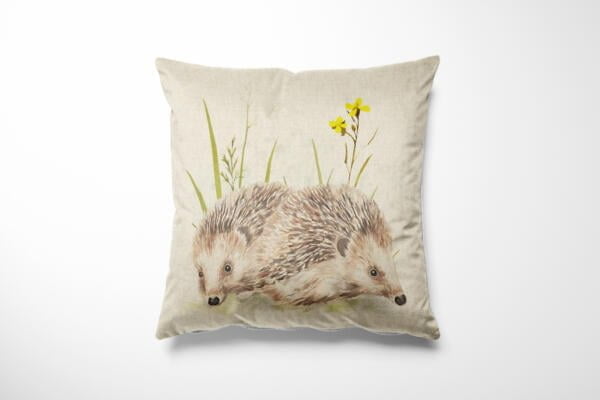 A square throw pillow with a beige fabric cover features a detailed illustration of two hedgehogs nestled together among tall grass and yellow flowers, creating a charming and nature-inspired decorative design.