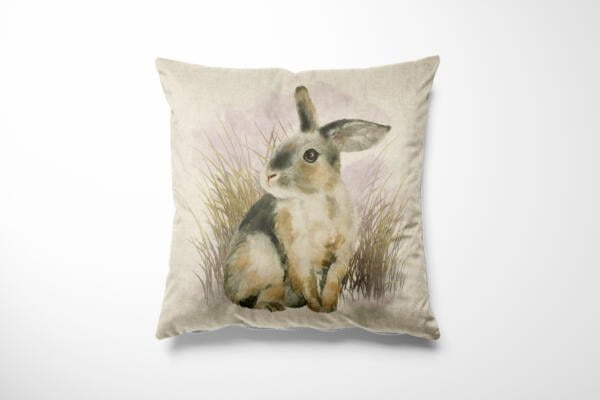 A decorative pillow with a watercolor painting of a rabbit. The rabbit has a mix of gray and white fur and is sitting in front of tall grasses on a light beige background.