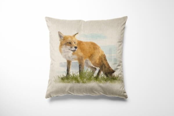 A beige cushion with a printed image of a red fox standing in grass. The fox is facing slightly to the side, showing its profile with a backdrop of a lightly colored, abstract sky. The cushion is placed on a white surface.