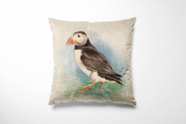 A decorative throw pillow featuring a detailed illustration of a puffin. The puffin is standing against a soft pastel background with shades of green and blue, creating a serene and natural setting. The pillow has a beige fabric and a square shape.
