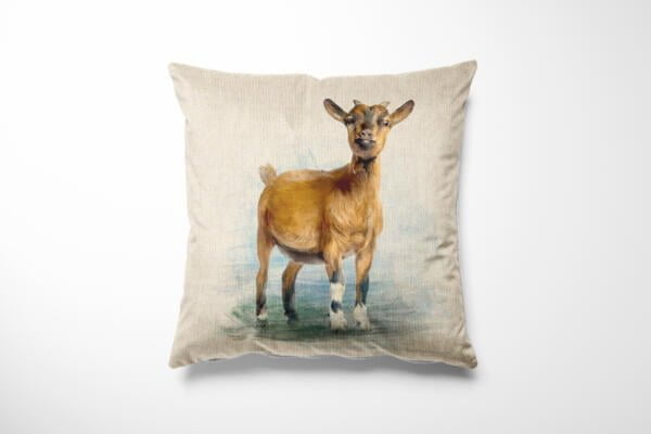A square beige throw pillow features a realistic illustration of a brown goat standing on a light background. The goat has short fur, pointy ears, and a prominent beard, with a slightly blurred meadow scene behind it.