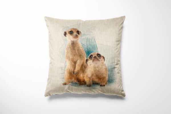 A beige throw pillow with an illustration of two meerkats. One meerkat is standing upright while the other is sitting beside it against a light blue abstract background. The pillow has a slightly textured fabric.