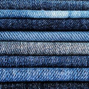 A close-up shot of a stack of Denim | Denim Fabric | Denim Fabric UK displaying various shades and textures, neatly folded and layered on top of each other. The denim fabric ranges in color from light blue to dark indigo, showcasing different patterns and weaves.